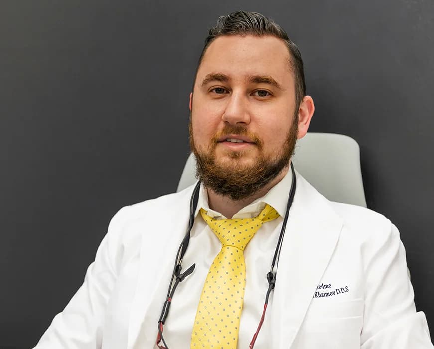 Dr. Khaimov in white lab coat and yellow tie sitting in chair with stethoscope around neck facing camera.