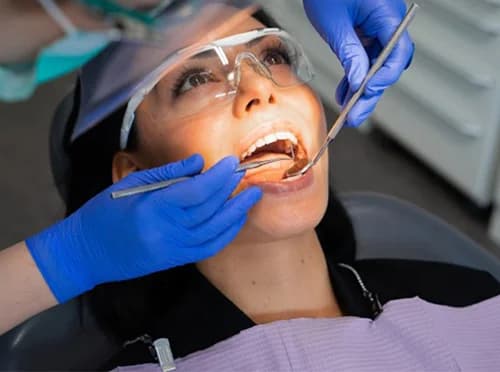 Female patient wearing safety glasses lying in a dental chair receiving dental implants Astoria dentist in blue gloves.