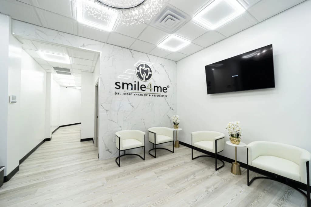Modern dental office waiting room with white chairs wood floor large TV on wall and Smile4Me logo with flowers on small tables.