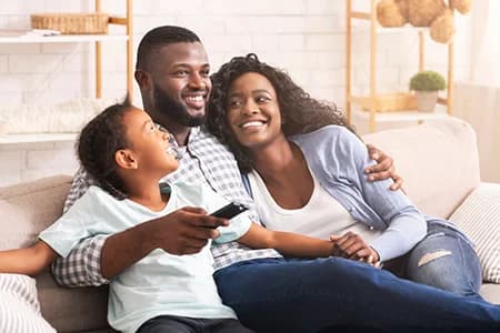 Smiling family sitting on couch father holding remote mother and child laughing together in bright living room with cozy decor.