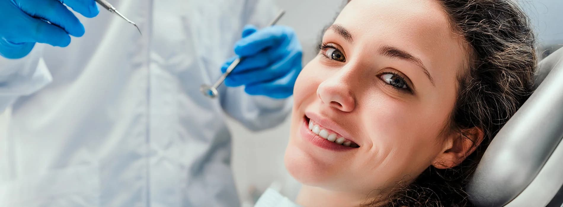Smiling woman lying in a dental chair looking at the camera while a dentist in white coat and blue gloves prepares dental tools in the background in a clean modern dental office.